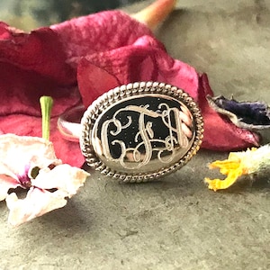 May include: A silver ring with an oval-shaped band and a raised, engraved monogram. The ring is set against a background of dried flower petals.