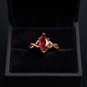 May include: A gold ring featuring a marquise-cut red gemstone set in a twisted band. The ring is displayed inside a black velvet-lined jewelry box, highlighting the contrast between the vibrant red stone and the polished yellow gold metal setting.