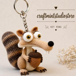 Può includere: Un portachiavi all'uncinetto a forma di scoiattolo beige con grandi occhi bianchi, naso nero e due denti bianchi, che tiene in mano una ghianda marrone. Lo scoiattolo ha una coda marrone a strisce ed è fissato a un anello in metallo argentato. Testo: craftmintstudiostore.