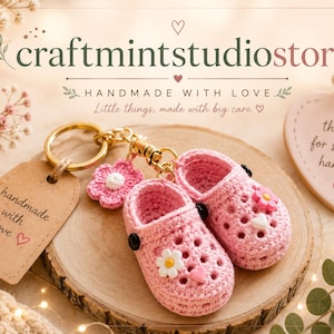 May include: A pair of miniature pink crocheted clogs attached to a gold-toned metal keychain. The clogs feature small white daisy and pink heart embellishments. A brown paper tag reads handmade with love, and a heart-shaped sign says thank you for supporting handmade.