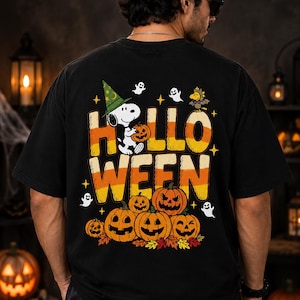 May include: A black short-sleeved t-shirt featuring a graphic on the back with the text "HALLOWEEN" in candy corn colours. The design includes Snoopy wearing a green witch hat, Woodstock, ghosts, stars, and a cluster of carved jack-o'-lanterns.