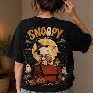 Może przedstawiać: Czarny t-shirt z grafiką na plecach przedstawiającą Snoopy'ego w czarodziejskim kapeluszu i pelerynie, siedzącego na czerwonej budzie z wiaderkiem w kształcie dyni. Wzór zawiera pełnię księżyca, duszki, latarnię oraz napis SNOOPY TRICK OR TREAT.