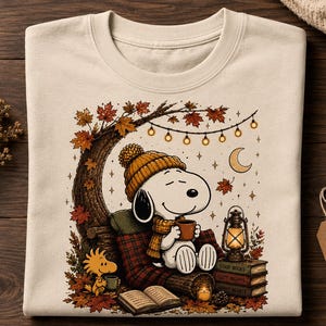 May include: A cream-colored crewneck sweatshirt featuring a graphic of a white beagle wearing an orange knit hat and scarf, sitting under a tree with autumn leaves, holding a mug, with a lantern, books, and a small yellow bird nearby.