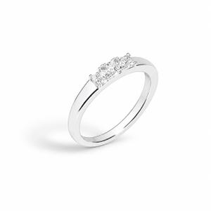 Könnte beinhalten: Ein polierter silberfarbener Ring mit einer Reihe von vier kleinen, klaren, rund geschliffenen Steinen, die horizontal oben eingesetzt sind. Die Ringschiene ist glatt und massiv mit einer hochglänzenden Oberfläche, die das Licht reflektiert.