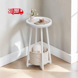 May include: A white round side table with three legs and a raised edge. On the tabletop sits a small vase with pink flowers, a book, and a cup. A beige woven basket is positioned between the legs on the lower level, containing soft items.