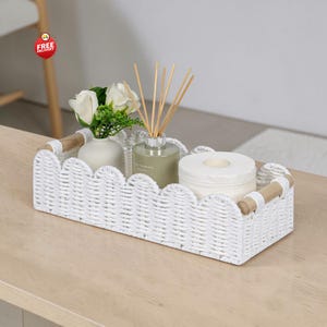 Op de afbeelding: Een rechthoekige witte gevlochten mand met een geschulpte bovenrand en lichte houten handvatten. In de mand staan een kleine witte vaas met witte kunstmatige rozen, een glazen geurstokjesdiffuser en een rol wit toiletpapier.