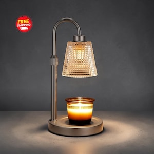 Puede incluir: Una lámpara calentadora de velas de metal cepillado con un brazo curvo y una pantalla de vidrio transparente texturizado. La lámpara ilumina una pequeña vela en frasco de vidrio ámbar sobre su base circular, creando un brillo cálido bajo la luz.