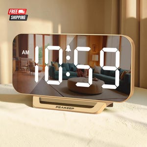 May include: A rectangular digital alarm clock with a reflective mirror surface and a light-colored base. The display shows the time 10:59 in bright white LED digits, including an AM indicator and a small bell icon. The brand name PEAKEEP is printed on the base.