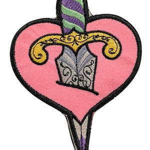 May include: An embroidered patch featuring a pink heart shape pierced by a dagger. The dagger has a gold-colored crossguard, a blade with intricate silver and black scrollwork, and a handle with alternating diagonal stripes of purple and light green.