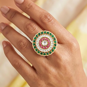 May include: A circular statement ring featuring a central clear stone surrounded by a ring of pink stones, an outer ring of green stones, and a border of small white beads. The metal setting is gold-toned. The text Gemoire is visible in the corner.