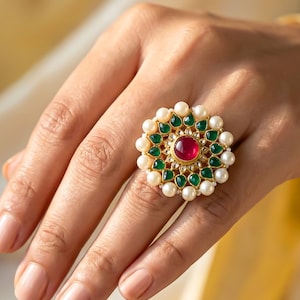 Può includere: Un anello color oro con una pietra rossa rotonda centrale circondata da una disposizione floreale di pietre verdi a goccia e un bordo di perle bianche. La parola Gemoire è stampata nell'angolo in alto a sinistra dell'immagine.