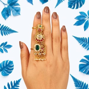 Op de afbeelding: Een goudkleurige ring voor meerdere vingers met gedetailleerd filigraan metaalwerk, een centrale ovale groene steen en decoratieve accenten in rood en wit. Het sieraad heeft kleine bungelende parelachtige kraaltjes en wordt over twee vingers gedragen.