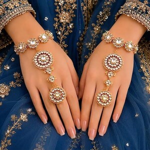 May include: A gold-toned hand harness jewelry set featuring circular motifs adorned with white stones and red accents. The design includes a bracelet connected by a delicate chain to a ring, resting against a deep blue fabric with gold embroidery.