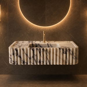 Peut inclure: Un lavabo rectangulaire mural en marbre blanc avec des veines gris foncé marquées. La vasque présente un design extérieur cannelé à nervures verticales distinctes et est équipée d'un robinet unique de couleur dorée orienté vers le haut.