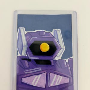 Puede incluir: Una tarjeta coleccionable rectangular dentro de una funda de plástico transparente que presenta una ilustración dibujada a mano de un robot morado con un ojo circular amarillo, componentes de cabeza angulares y una placa pectoral con dos triángulos.