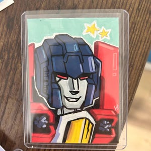 Peut inclure: Une carte à collectionner rectangulaire présentant une illustration dessinée à la main d'un personnage robot avec un casque bleu foncé, des yeux rouges et un visage argenté. Le robot a des épaulettes rouges et une plaque de poitrine jaune sur fond bleu clair.