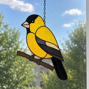 May include: A stained glass suncatcher shaped like a goldfinch with bright yellow, black, and white glass panels. The bird is perched on a brown branch and hangs from a silver metal chain, featuring dark lead lines outlining each glass segment.
