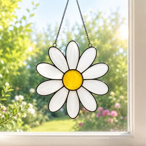 Può includere: Un acchiappasole in vetro colorato a forma di fiore di margherita con dodici petali bianchi e un centro giallo brillante. Il pezzo è incorniciato da linee di saldatura in metallo nero e pende da una sottile catena di metallo nero davanti a una finestra.