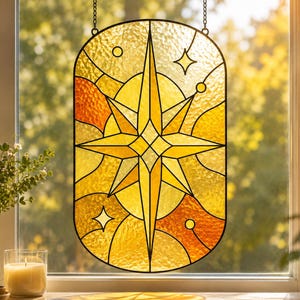 Può includere: Un acchiappasole in vetro colorato di forma ovale con una stella centrale a otto punte in tonalità gialle e arancioni. Il design include linee geometriche, piccoli cerchi e stelle a quattro punte, il tutto racchiuso in una cornice di metallo scuro.