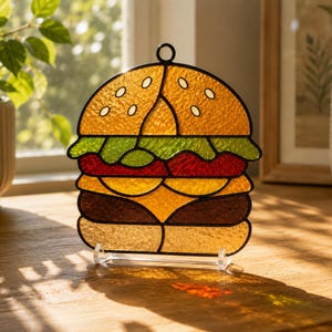 Può includere: Un acchiappasole in vetro colorato a forma di hamburger con un panino dorato con semi di sesamo bianchi, lattuga verde, una fetta di pomodoro rosso, formaggio giallo e una polpetta marrone, il tutto bordato di metallo nero su supporto trasparente.