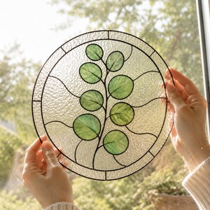 Puede incluir: Un atrapasoles circular de vidrieras con un diseño central de rama con hojas verdes redondas. La pieza está construida con paneles de vidrio transparente texturizado unidos por líneas de plomo de metal oscuro, creando un adorno botánico para ventanas.