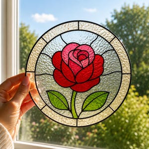 Può includere: Un acchiappasole circolare in vetro colorato con una rosa rossa centrale con centro rosa e due foglie verdi. Il design è racchiuso in una cornice di piombo nera con uno sfondo in vetro trasparente testurizzato, come ornamento per finestra.