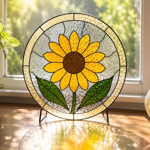 Può includere: Un pannello circolare in vetro colorato raffigurante un girasole giallo centrale con un cuore marrone e due foglie verdi. Il fiore è posto su uno sfondo di vetro trasparente testurizzato con linee di piombo nere, sostenuto da un supporto in metallo nero.