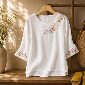 May include: A white blouse with three-quarter length sleeves and a V-neckline. The garment features delicate floral embroidery in shades of peach and green around the neckline and on the sleeve cuffs. It includes small white button accents.