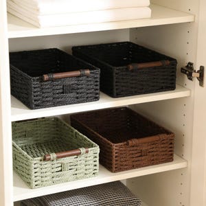 May include: Four rectangular storage baskets with a woven texture and dark brown wooden handles. Two baskets are black, one is dark brown, and one is sage green. The baskets are arranged on two white shelves inside a cabinet with white folded linens.