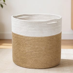 以下が含まれることがあります： 巻かれたロープで作られた円筒形の収納バスケット。上部は白、下部は自然なベージュ色です。バスケットの上部の縁には2つの統合されたハンドルが切り込まれており、構造化されたデザインを提供しています。