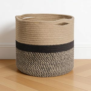 May include: A cylindrical storage basket constructed from coiled rope. The upper section is a solid tan colour, while the lower section features a black and tan diagonal striped pattern. The basket includes two integrated handles at the top rim.