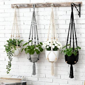 May include: Four macrame plant hangers in cream, grey, and black, each featuring knotted cord designs, wooden or plastic beads, and a bottom tassel. These hangers support various potted plants, including a trailing vine and white flowers.