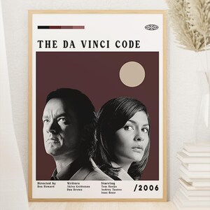 May include: A framed film poster for The Da Vinci Code featuring black and white portraits of two people against a dark maroon background with a beige circle. Text includes credits for director Ron Howard, writers Akiva Goldsman and Dan Brown, stars Tom Hanks, Audrey Tautou, and Jean Reno, and the year 2006.