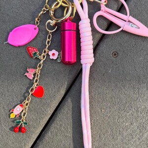 May include: A gold-toned carabiner keyring featuring a pink cylindrical pill case, a long pink paracord lanyard, and a gold chain with charms including a strawberry, a pink flower, a butterfly, a red heart, a rocket, and cherries, plus pink scissors.