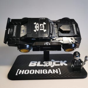 Peut inclure: Un modèle de voiture LEGO Ford Hoonigan noir avec des roues dorées, arborant un logo tête de mort, le numéro 43 et la marque Ford. Il est présenté sur un support noir marqué BLOCK HOONIGAN, accompagné d'une figurine de pilote de course assortie.