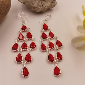 May include: A pair of chandelier earrings featuring multiple faceted, teardrop-shaped red stones set in silver-toned metal. Each earring consists of several tiers of these red gems connected by small silver rings, suspended from a silver hook wire.