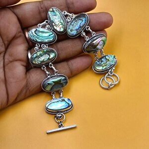 Peut inclure: Un bracelet en argent composé de sept cabochons ovales en coquille d'ormeau aux reflets irisés verts, bleus et violets. Chaque coquille est sertie dans un cadre en argent torsadé et reliée par des maillons, avec un fermoir à barre et anneau.