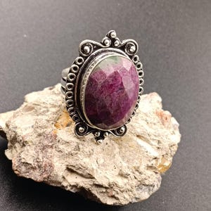 Puede incluir: Un anillo de plata con una gran piedra ovalada facetada de color rubí, engastada en un marco de metal decorativo con intrincados adornos de volutas y pequeños detalles circulares que rodean la gema central, con un acabado pulido y texturizado.