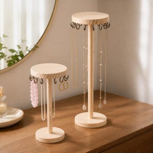 May include: Two light-coloured wooden jewellery stands with circular bases and tops. Each stand features multiple dark metal hooks along the underside of the top disc, holding various necklaces, a pink beaded bracelet, and gold hoop earrings.