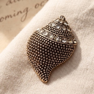 May include: A seashell-shaped brooch with a textured, antiqued gold-tone finish. The upper spiral section is adorned with a row of clear, round-cut rhinestones, while the lower body features a dense, stippled pattern across the surface.