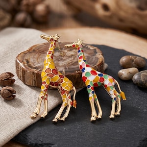 May include: Two giraffe-shaped brooches with gold-tone metal outlines. One brooch features yellow and orange enamel spots with a glittery finish. The second brooch displays a multicoloured pattern of red, blue, green, and white enamel spots across its body.