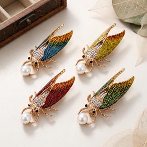May include: Four cicada-shaped brooches in gold-tone metal with enamel wings in blue, yellow, red, and green. Each insect features clear rhinestones on the thorax, black bead eyes, and a single white faux pearl held by the front legs.