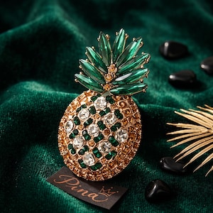 Op de afbeelding: Een ananasvormige broche met een goudkleurige metalen basis ingelegd met heldere, amberkleurige en donkergroene strass-steentjes. De bovenste bladeren bestaan uit langwerpige donkergroene kristallen. Een zwart label toont het woord Pina en een kroon.