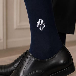 May include: A pair of navy blue crew-length socks featuring a white embroidered monogram with the letters ABK in a geometric font. The socks are worn with black leather cap-toe oxford shoes and dark trousers, showcasing a ribbed cuff design.