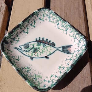 May include: A square ceramic dish with rounded corners, featuring a white base with a green speckled pattern. The centre of the dish displays a stylised line drawing of a fish with dark outlines, green interior speckles, and red accents on the fins.