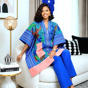 May include: A vibrant blue tunic featuring a multi-coloured floral pattern, vertical stripes, and solid peach-coloured pockets. The garment has a V-neckline and wide sleeves, paired with matching blue trousers and a large, matching blue headpiece.