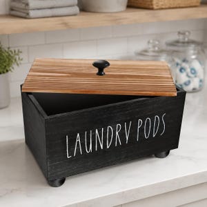 May include: A rectangular black wooden storage box with a natural wood grain lid and a small black knob. The front of the box features the white text LAUNDRY PODS in a handwritten style font. The container stands on four small black feet.