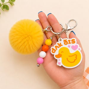 May include: A keychain featuring a large, round, yellow faux fur pom-pom, a strand of four colorful silicone beads in yellow, orange, white, and pink, and an acrylic charm shaped like a yellow rubber duck with a pink heart and the text "duck dis".