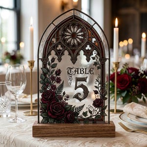 May include: A stained glass table number sign featuring the text "Table 5" in a gothic font. The arched glass panel is decorated with dark red roses and green foliage, framed in black metal, and set into a rectangular wooden base.