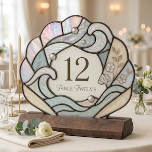 May include: A stained glass table number sign shaped like a seashell with iridescent pastel panels, featuring the number 12 and the text Table Twelve in the center. The piece is set in a dark wooden base and decorated with small glass spheres.